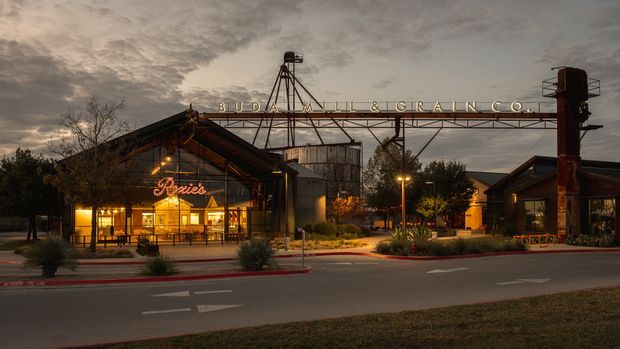 Twilight exterior view of a repurposed industrial-style commercial complex with a prominent steel grain-elevator structure, weathered metal buildings, exposed roof framing, warm glowing signage, landscaped planting, and a street frontage. The elevated sign reads “Buda Mill & Grain Co.”