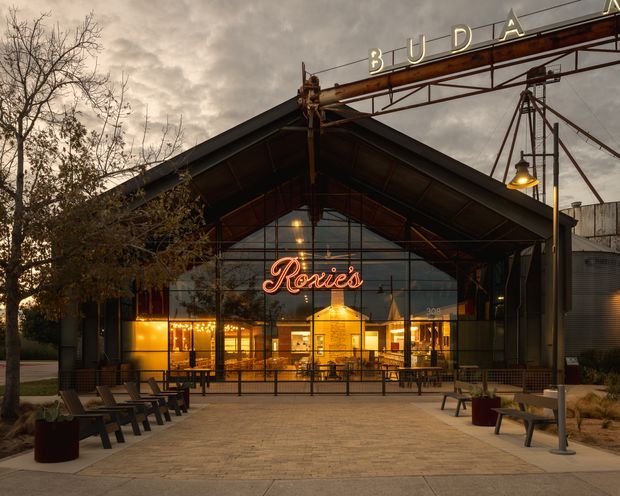 Exterior view of a large industrial-style hospitality or commercial venue at dusk. The building has a dramatic pitched steel canopy, extensive floor-to-ceiling glazed frontage, exposed structural steel, an illuminated neon “Rosie’s” sign, outdoor bench seating, and a prominent overhead “Buda” sign structure. Warm interior lighting contrasts with the dark metal facade and overcast sky.