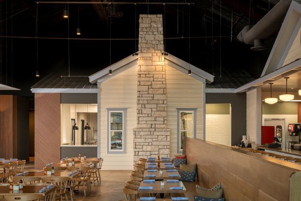 Interior view of a warmly lit restaurant or hospitality space with a prominent small gabled cottage-like structure, pale weatherboard-style walls, and a central full-height light stone chimney. The room has a dark industrial ceiling with exposed services and suspended pendant lights, timber dining tables and chairs, a long upholstered banquette, and a service counter with drinks equipment.