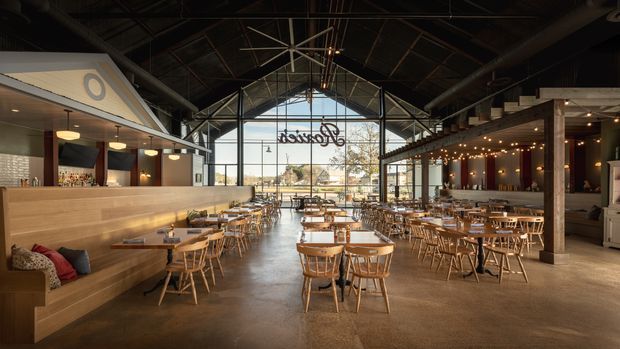 A spacious industrial-style hospitality interior, likely a restaurant or brewery, with a high exposed dark steel roof structure, large floor-to-ceiling glazed gable end, polished concrete flooring, extensive timber dining tables and chairs, a long built-in timber banquette, and bar/service counters along both sides. Warm pendant lighting and festoon lights create an inviting dining atmosphere.