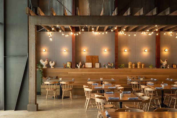 A warm, rustic-industrial dining space with rows of wooden tables and chairs, a long timber banquette, decorative shelving, exposed structural framing, corrugated metal upper walls, and string lights. The room has a tall mezzanine-like upper level and a muted palette of timber brown, grey, and warm cream.