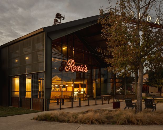 Street-level exterior view of a contemporary industrial-style hospitality or commercial building at dusk. The facade combines dark metal framing, expansive glazed frontage, translucent or perforated metal screening, and a projecting pitched canopy. Warm interior lighting reveals a restaurant or bar with a prominent neon “Rosie’s” sign, outdoor seating, pedestrian paving, and landscaped ornamental grasses and trees in the foreground.