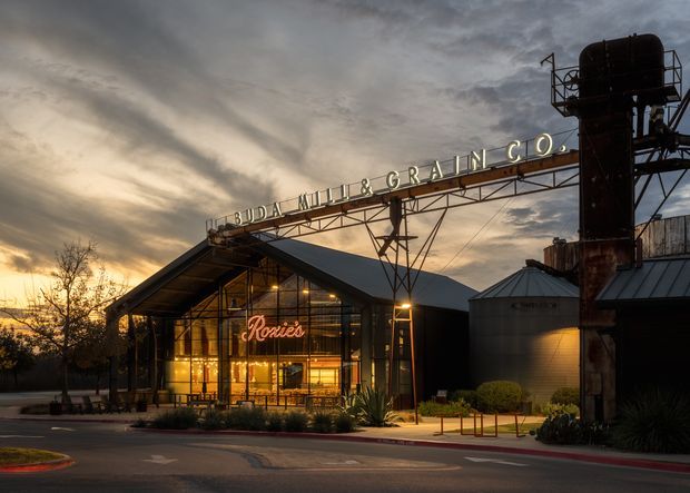Exterior view of the Ruda Mill & Grain Co. building at dusk, featuring a restored industrial-style grain facility with a prominent steel gantry sign, corrugated metal roofs, grain silos, elevated machinery, and a warmly illuminated glazed restaurant or commercial frontage.