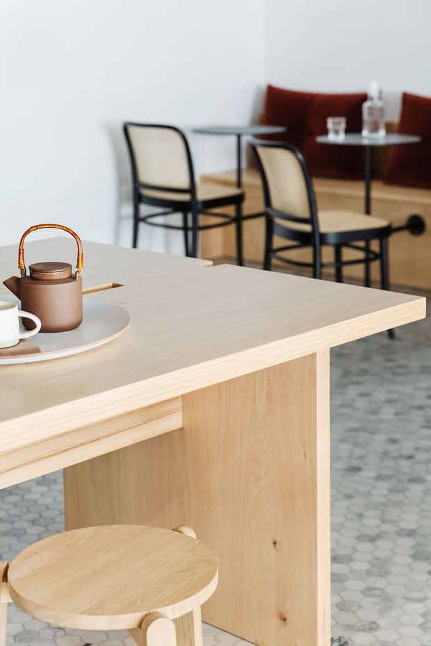 A softly lit contemporary café or hospitality interior featuring a pale timber counter with integrated panel detailing and a round timber stool in the foreground. A tea set rests on a tray atop the counter. In the background are small round café tables, black-framed woven chairs, and a built-in tan leather banquette. The floor is finished with small light grey hexagonal tiles, while the walls are plain white.