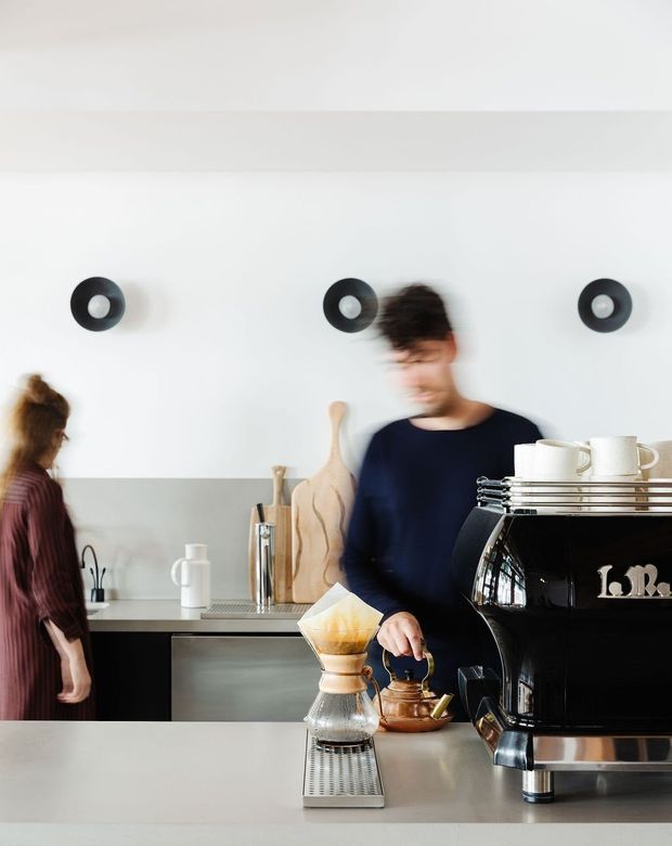 A bright, modern café or commercial coffee bar interior with a large professional espresso machine in the foreground, pour-over coffee equipment on the counter, and two people working behind a sleek preparation counter. The space features white walls, dark lower cabinetry, stainless-steel surfaces, and simple black circular wall lights.