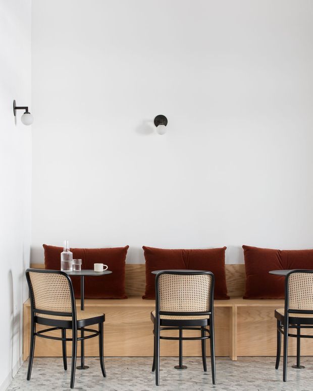 Minimalist café or hospitality interior with a long built-in timber banquette against a white wall, upholstered in deep rust-red cushions. Black-framed cane dining chairs and small round black tables face the seating. The space has a pale terrazzo floor, simple wall-mounted globe lights, and a restrained contemporary composition.
