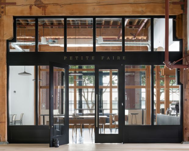 Interior view toward a black-framed glazed entrance labelled “PETITE FAIRE,” with timber wall and ceiling structure, exposed services, industrial pendant lighting, and a bright café or commercial workspace visible beyond the doors.