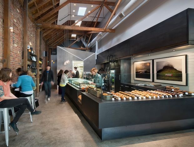 A long, narrow bakery or café interior in a converted industrial-style space. The room features exposed red brick walls, a high timber roof structure with exposed beams and rafters, polished concrete flooring, and a large black service counter with integrated display cases and cabinetry. Customers sit along a bar at the left, while staff work behind the counter. Pendant and recessed lighting, framed artwork, and a glazed preparation area complete the commercial fit-out.