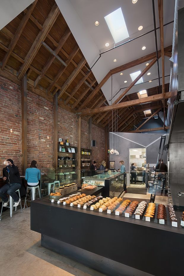 Interior view of a bakery or café in a converted industrial space. The room features exposed red-brick walls, a dramatic exposed timber roof structure with rafters and beams, polished concrete flooring, black commercial display counters, pendant and recessed lighting, and multiple roof skylights. Pastries are arranged in glass display cases, with customers seated along the left wall and a preparation area visible at the rear.