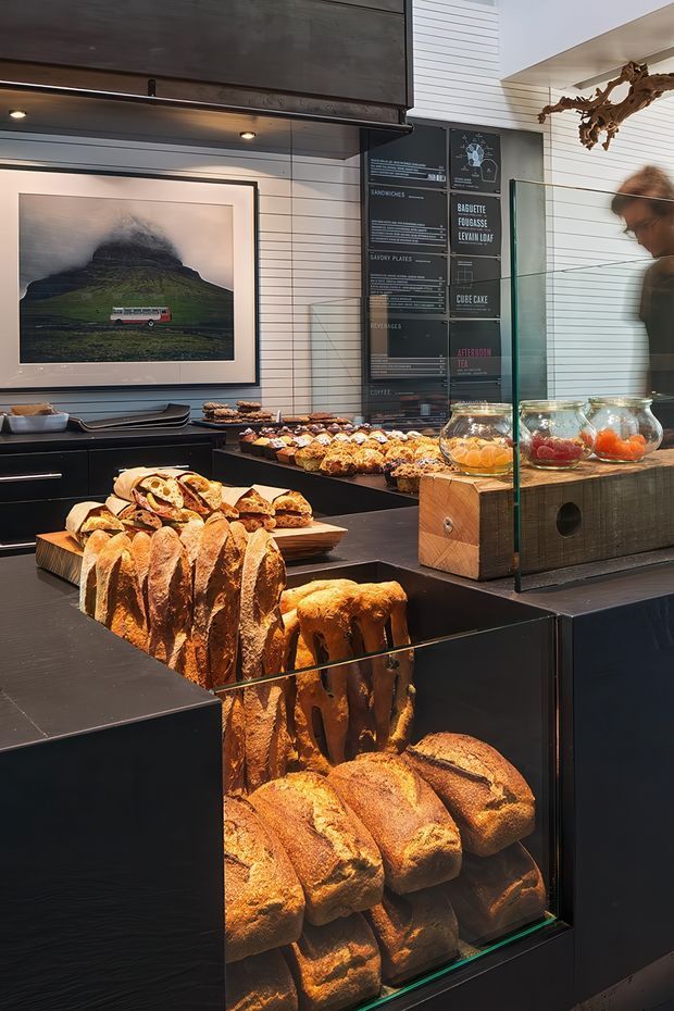 Interior view of a contemporary bakery or café retail counter, with extensive bread and pastry displays arranged on dark built-in cabinetry and behind a glass display screen. The space features black counters, white horizontal wall cladding, menu boards, recessed under-counter lighting, and a framed landscape photograph. A timber display box and glass ingredient jars add warmth to the otherwise dark, refined fit-out.