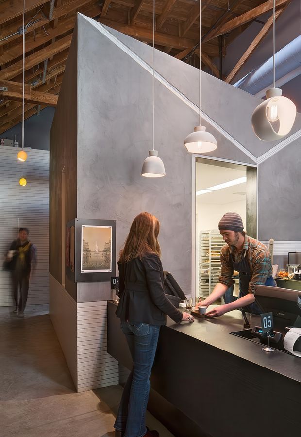 Interior photograph of a contemporary commercial café or food-service counter. A customer is being served at a dark counter with point-of-sale terminals, while a staff member works behind it. The space features a dramatic exposed timber roof structure, large grey concrete-look partition walls, suspended pendant lights, visible metal ductwork, and corrugated metal wall lining. The image has a warm industrial character with active service operations.