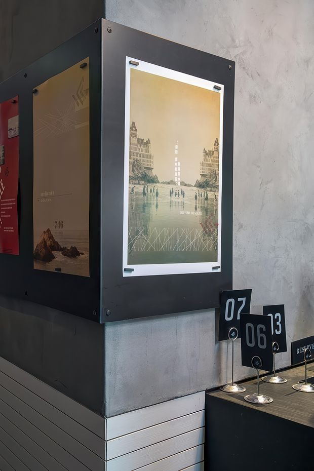 A close-up view of a dark, industrial-style exhibition or hospitality interior. Black metal display panels hold vintage travel-style posters mounted against a textured concrete wall, while a dark counter in the foreground displays numbered table or reservation markers. The space has a moody, graphic, utilitarian character with concrete, metal, and black finishes.