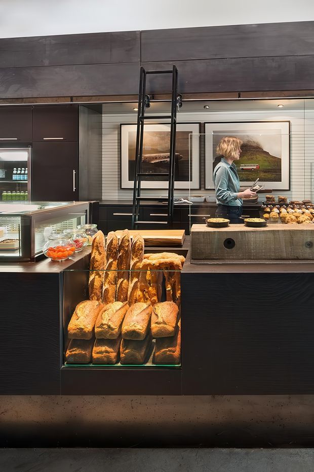 A dark, sophisticated bakery or café interior with extensive black cabinetry and counters, illuminated glass bread display, refrigerated shelving, and a central timber-toned service counter displaying pastries. A black rolling ladder reaches tall overhead storage, while framed artwork and horizontal wall panelling add visual interest. Warm task and display lighting contrasts with the charcoal finishes.