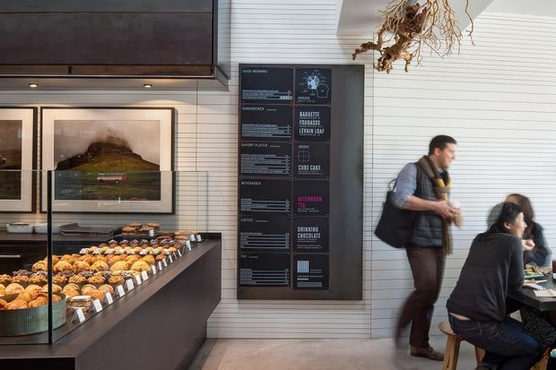 A bright, contemporary café or bakery interior with a long dark service counter displaying rows of pastries behind glass. The space features white horizontal wall cladding, framed artwork, a large black menu board, dark cabinetry, and customers seated at a small table. A hanging dried botanical installation adds a natural accent.