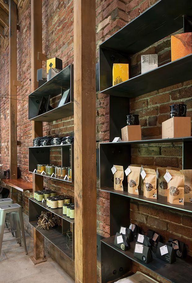A narrow retail interior with exposed red brick walls, substantial vertical timber posts, and black metal display shelving arranged in stepped geometric modules. The shelves hold packaged coffee or specialty food products, bottles, tins, and boxed goods. A concrete floor, industrial shelving, and exposed masonry create a warm warehouse-style shop atmosphere.
