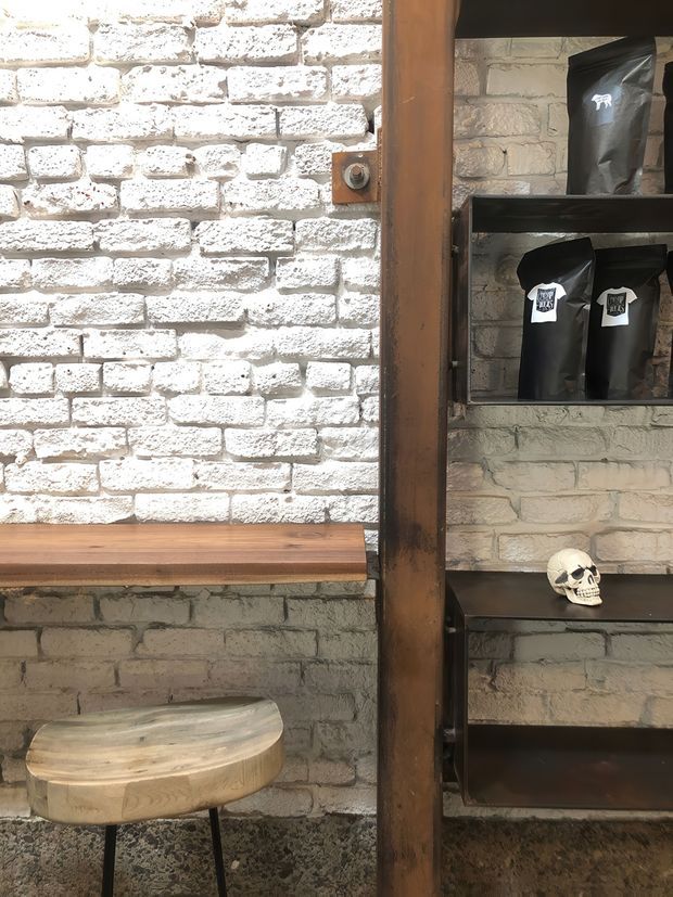 A rustic industrial commercial interior corner with whitewashed exposed brick walls, dark metal open shelving displaying packaged goods and a skull ornament, a thick timber structural post, a timber wall-mounted bench or counter, and a small round wooden stool. The space has a warm, moody, textural atmosphere with strong industrial detailing.