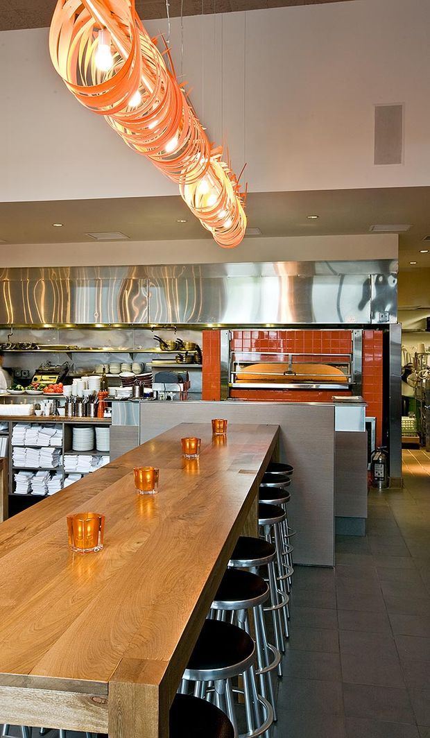 Interior view of a contemporary commercial restaurant, focused on a long solid timber communal dining table with black stools and small candle holders. Behind it is an open professional kitchen with stainless-steel exhaust cladding, shelving, cooking equipment, and a red ceramic-tiled pizza oven. A large sculptural orange pendant light runs diagonally across the ceiling. The space combines warm timber and orange lighting with cool stainless steel and dark tiled flooring.