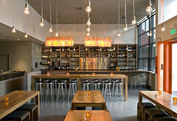 A contemporary commercial bar and dining space with a long timber communal table, stools, built-in bar counter, extensive back-bar shelving stocked with bottles, and a row of beer taps. Numerous exposed pendant bulbs and two bands of warm illuminated feature lighting hang above the bar. Large black-framed windows and an orange-framed glazed entrance provide strong daylight, while dark polished flooring and neutral walls create an industrial urban atmosphere.
