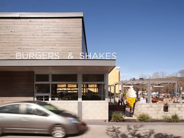 Exterior street view of a contemporary fast-food restaurant with a simple rectangular form, horizontal timber cladding, broad roof overhang, storefront glazing, and prominent “BURGERS & SHAKES” signage. A covered outdoor dining area and ice-cream sign are visible beside the building, with a car passing in the foreground.