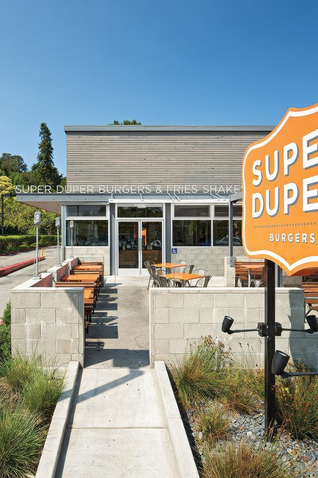 Exterior street-level view of a contemporary burger restaurant with a low flat roof, horizontal timber-clad upper facade, pale concrete block walls, extensive storefront glazing, and a landscaped outdoor dining terrace. Orange branded signage dominates the right foreground, while concrete pathways, timber tables, chairs, and grasses define the approach.