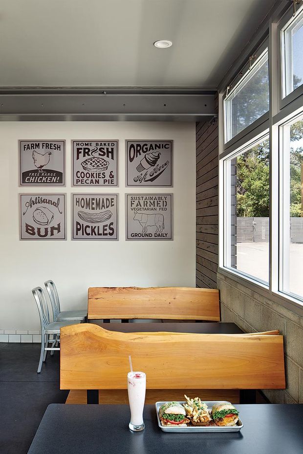 A compact casual dining space with built-in timber booth seating, dark tables and chairs, large black-framed windows, and a charcoal tiled floor. The walls feature a collection of vintage-style food and farm-themed posters. Exposed dark timber wall cladding and visible metal framing give the room a simple contemporary-industrial character, with abundant natural light from the glazing.