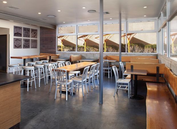 A bright contemporary café or casual dining space with extensive glazing, timber banquette seating, communal timber tables, mixed metal and timber chairs, and polished dark concrete flooring. Slim structural columns divide the open room, while framed graphic wall art and timber wall panels add character. Outdoor shade structures and landscaping are visible beyond the windows.