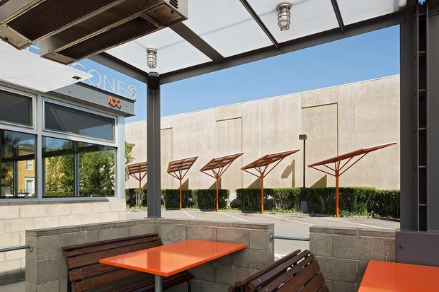 A covered outdoor courtyard or terrace seating area beside a commercial-looking building. The space has built-in grey masonry bench enclosures, dark timber slatted benches, bright orange tables, and a translucent roof canopy supported by dark metal framing. Beyond the seating is a paved forecourt with landscaped hedges and distinctive angular orange shade structures attached to a pale rendered facade. Large glazed windows and a partial building sign are visible at left.