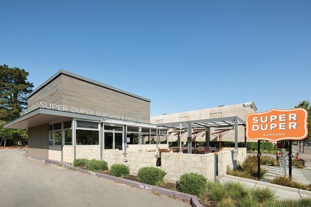Exterior view of a Super Duper Burgers restaurant, showing a low-rise contemporary commercial building with a tall angled roof volume, horizontal brick-like cladding, extensive glazing, and a covered outdoor dining terrace. A prominent orange restaurant sign stands in the foreground, with landscaped planting around the paved site.