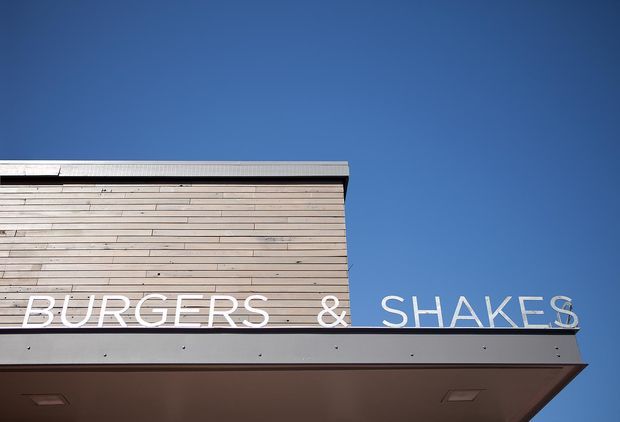Close-up exterior view of a contemporary commercial building facade under a clear blue sky. The facade combines horizontal light timber-like cladding with a dark grey fascia and projecting canopy, featuring large white lettering reading “BURGERS & SHAKES.”