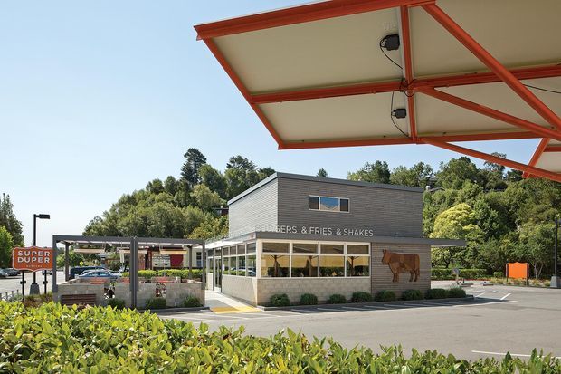 Exterior view of a single-storey Super Duper Burgers restaurant in a landscaped suburban setting. The building has a low, angular roof and a dark horizontal-cladding facade, extensive glazed dining-area windows, and prominent red-painted steel canopy framing extending over the forecourt. Restaurant signage and parking areas are visible.