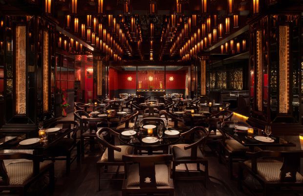 A dramatic, highly symmetrical upscale restaurant dining room with rows of dark timber tables and woven-seat chairs arranged beneath a dense grid of warm pendant lights. The space features glossy dark finishes, ornate framed wall detailing, red feature walls and screens, reflective surfaces, and intimate candlelit table settings, creating an opulent, theatrical atmosphere.
