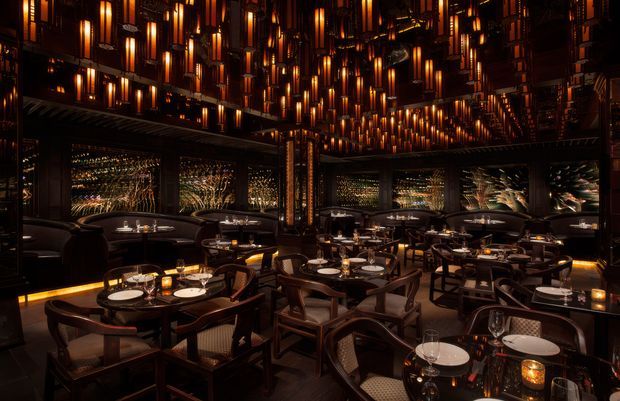 A dark, luxurious restaurant dining room filled with closely arranged tables, upholstered and timber-framed dining chairs, place settings, candles, and reflective decorative wall panels. Hundreds of warm amber pendant lights create a dramatic illuminated ceiling, while dark timber and metallic finishes produce an intimate, high-end atmosphere.