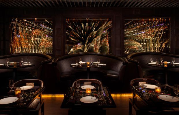 A dark, luxurious restaurant dining interior with several intimate tables arranged in front of curved banquette seating. The room features highly reflective mirrored wall panels with ornate, radiating decorative patterns, dark timber detailing, black glossy tables, candlelight, and warm amber floor lighting. A slatted dark ceiling reinforces the dramatic, richly layered atmosphere.