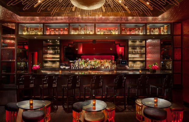 A richly detailed, dimly lit cocktail bar with a long dark bar counter, extensive illuminated bottle and glass shelving, red backlighting, ornate patterned wall finishes, and small round tables with stools in the foreground. A dramatic sculptural ceiling light creates a warm, glamorous atmosphere.