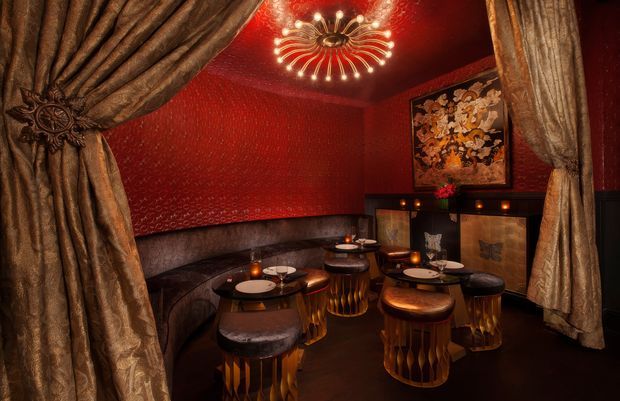 A richly decorated, intimate lounge or dining space framed by heavy metallic drapes. Deep red patterned walls, dark wainscoting, curved upholstered banquette seating, small round tables, and sculptural stools create a dramatic, theatrical atmosphere. Warm candlelight and an ornate radial chandelier provide accent lighting, while a framed artwork decorates the wall.