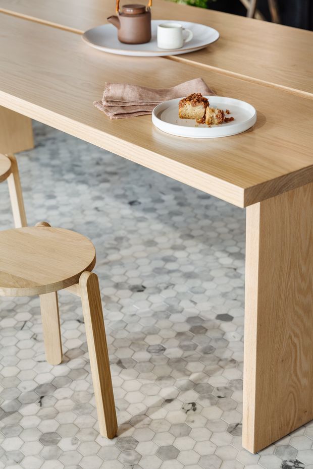 Close-up interior view of a light timber breakfast bar or kitchen island with a thick wood benchtop and matching wood-paneled support. A small round timber stool sits beside it. The floor is finished in a pale grey and white hexagonal tile pattern, while ceramic plates, cups, a teapot, folded napkin and pastry create a styled dining vignette.