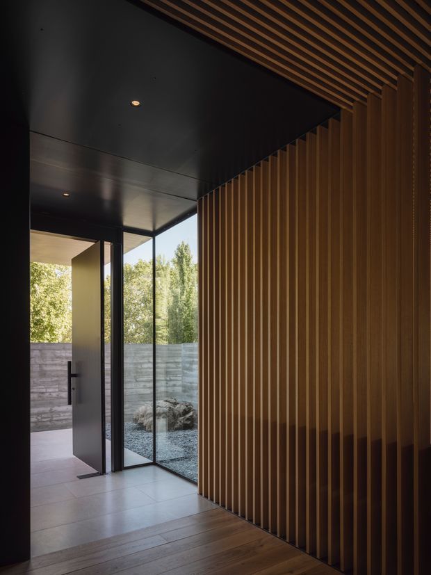 A dark, minimalist architectural entry space with a dramatic full-height vertical timber batten wall and matching slatted ceiling. A partially open black-framed glass door leads to a small gravel courtyard, while large-format pale floor tiles transition to warm timber flooring. Recessed lighting and strong light-and-shadow contrast emphasize the contemporary detailing.