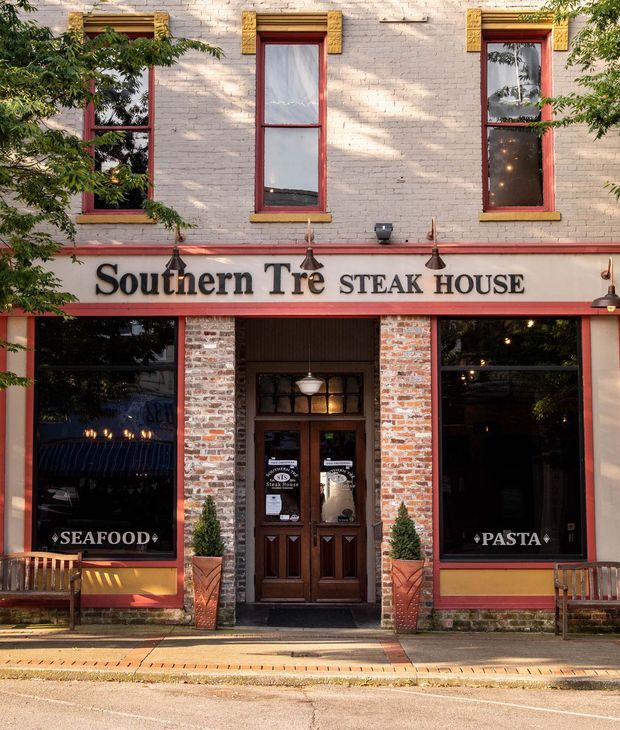 Street-facing historic brick restaurant facade for Southern Tré Steak House. The two-storey building has pale painted brick walls, tall red-painted timber window and door frames, decorative gold-toned window surrounds, red horizontal trim, exposed red brick around the ground-floor entrance, large dark shopfront windows, and prominent restaurant signage. Trees partially frame the facade, with a sidewalk and street in front.