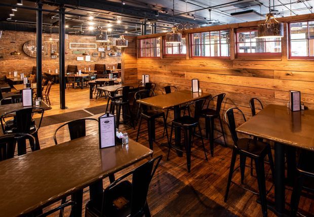 Interior view of a spacious rustic-industrial restaurant dining area with numerous dark wood tables, black metal chairs, exposed brick walls, timber wall panelling, and a dark timber floor. Large black-framed glass partitions reflect additional seating areas, while exposed ceiling services, track lighting, and woven pendant lights reinforce the industrial character.
