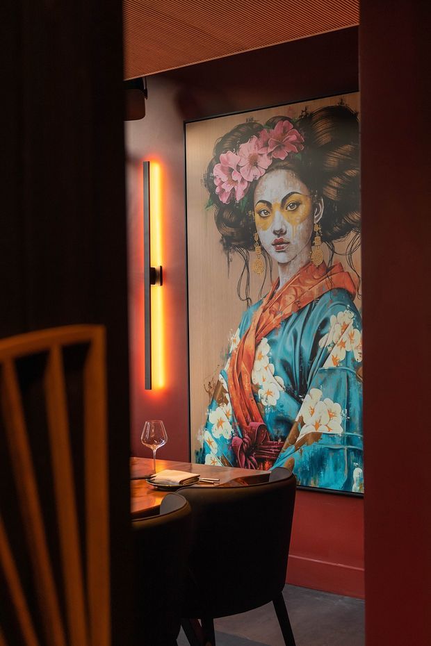 A moody, intimate dining area viewed through a dark doorway, featuring a round dining table with place settings, upholstered dark chairs, a large framed portrait artwork, warm vertical accent lighting, dark red walls, and a slatted ceiling detail.