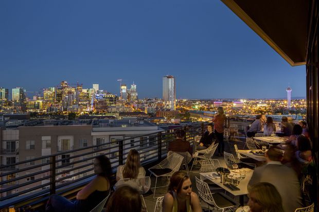 A busy rooftop terrace or balcony restaurant at dusk, furnished with outdoor tables and wire-frame chairs beside a dark metal railing. Diners occupy the seating area, while a brightly illuminated high-rise city skyline stretches across the background, including a prominent observation tower. The scene has a lively urban hospitality atmosphere with warm artificial lighting and deep blue evening sky.
