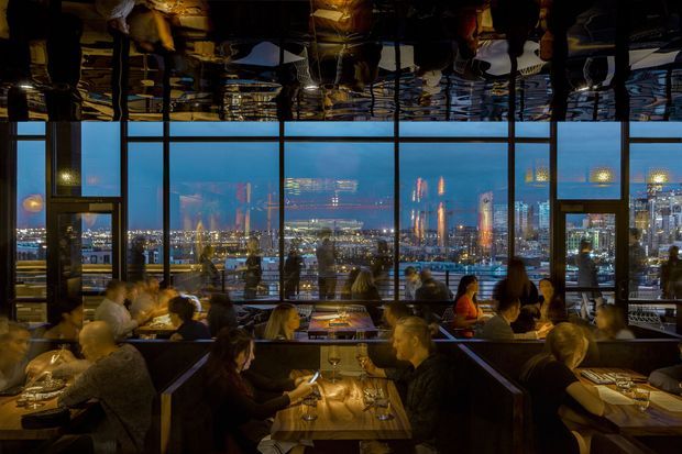 A dimly lit upscale restaurant dining room at dusk, with numerous diners seated in dark upholstered booths and tables arranged beside expansive floor-to-ceiling windows. The glazing provides a panoramic urban waterfront or harbour-city view with illuminated buildings and reflections, while the dark glossy ceiling mirrors the room below. Warm pendant and table lighting creates a sophisticated evening atmosphere.