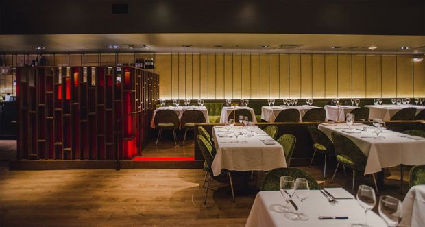 A dimly lit upscale restaurant dining room with neatly set tables, white tablecloths, wine glasses, upholstered chairs, warm timber flooring, and a decorative dark timber room divider accented with red lighting. The space has a refined, intimate atmosphere with linear wall panelling and concealed ceiling lighting.