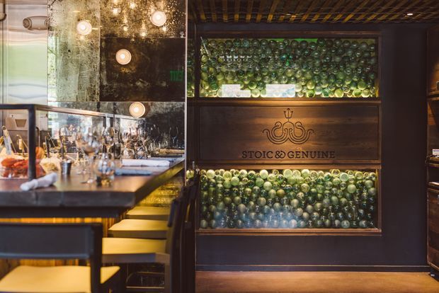 A dimly lit, richly detailed bar or restaurant interior featuring a polished bar counter with stools, mirrored wall surfaces, pendant and recessed lighting, and a prominent dark timber display wall. Backlit glass shelving filled with green and clear bottles frames a carved sign reading “Stoic & Genuine,” creating a sophisticated, atmospheric hospitality setting.