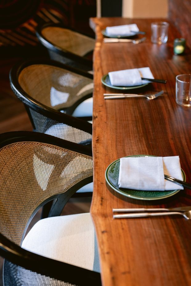 Close-up view of a warmly lit restaurant dining setting, with a long polished timber table set with green ceramic plates, folded white napkins, cutlery and drinking glasses. Black-framed woven cane dining chairs line the table, creating a refined yet natural hospitality interior.