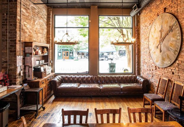 A rustic industrial-style commercial interior, likely a café, bar, or lounge, with exposed red-brick walls, tall street-facing windows, timber flooring, a large tufted brown leather sofa, wooden dining chairs and tables, vintage shelving, and exposed metal ductwork. A large round wall clock and pendant fixtures add character. The space is warmly lit with a strong blend of natural daylight and warm timber and leather tones.