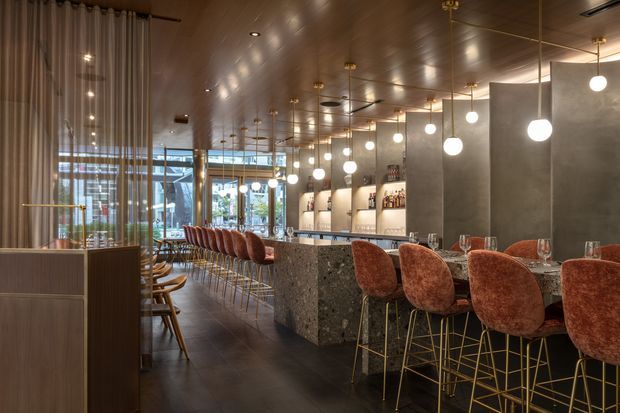 A sophisticated commercial bar and dining interior with a long stone island counter, rows of rust-coloured upholstered bar stools on slender gold-toned frames, and a second seating area beside floor-to-ceiling curtains. The space features dark tiled flooring, timber-toned ceiling panelling, concrete-look partition walls, illuminated shelving with bottles, and many suspended globe pendant lights. Glazed frontage brings in daylight, creating a warm, refined hospitality atmosphere.