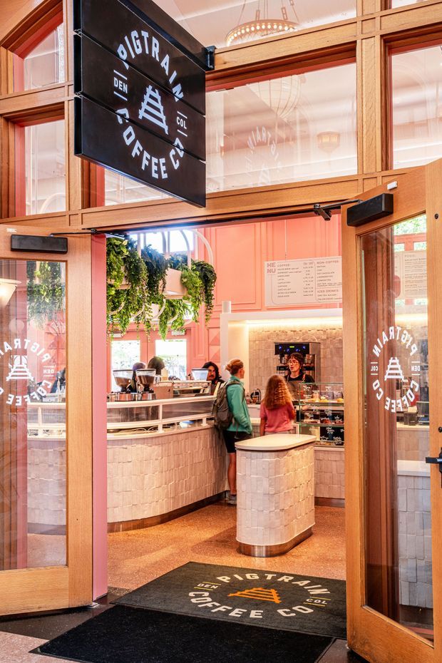 View through open timber-framed glass doors into a brightly styled coffee shop. The interior features coral-pink wall panelling, warm timber framing, a tiled service counter and central pedestal, hanging greenery, illuminated menu and coffee equipment, branded glazing decals, and a black entrance mat. The space has a polished hospitality fit-out with strong warm tones and abundant natural light.