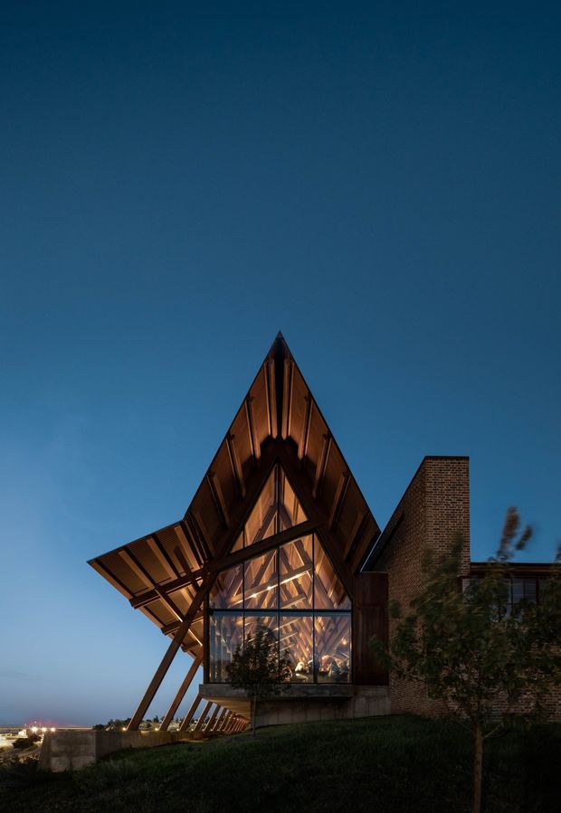 Dusk exterior view of a dramatic contemporary building with a steep triangular, gabled timber structure and expansive glazed facade. Warm interior lighting reveals exposed timber framing and rafters, while a projecting wing is supported by angled structural posts. A brick chimney and adjoining brick volume contrast with the timber and glass. The building sits on a gently sloping landscaped site beneath a deep blue evening sky.