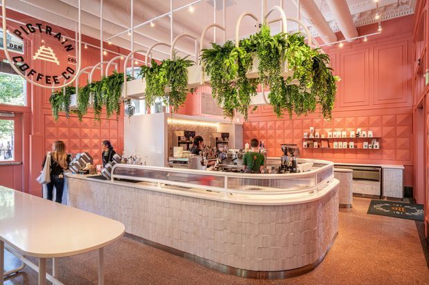 A bright, highly styled coffee shop interior dominated by a large curved central service counter. The counter has pale textured tile cladding and a light benchtop, with coffee equipment and staff behind it. The walls are painted coral pink with decorative panel moulding and geometric relief tiles, while numerous hanging plants are suspended from a white exposed ceiling structure. A large circular coffee-brand sign, display shelving, built-in cabinetry, tables, and stools complete the commercial café setting.
